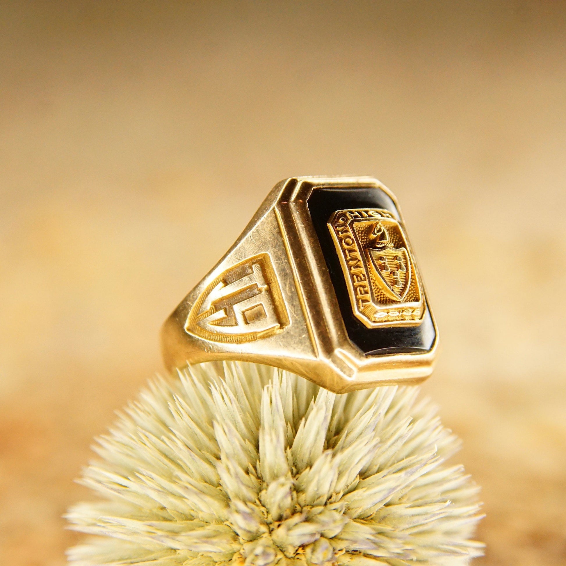 Vintage 1938 10K yellow gold and onyx Trenton High School class ring with engraving, sitting on a dried white flower, against a tan background