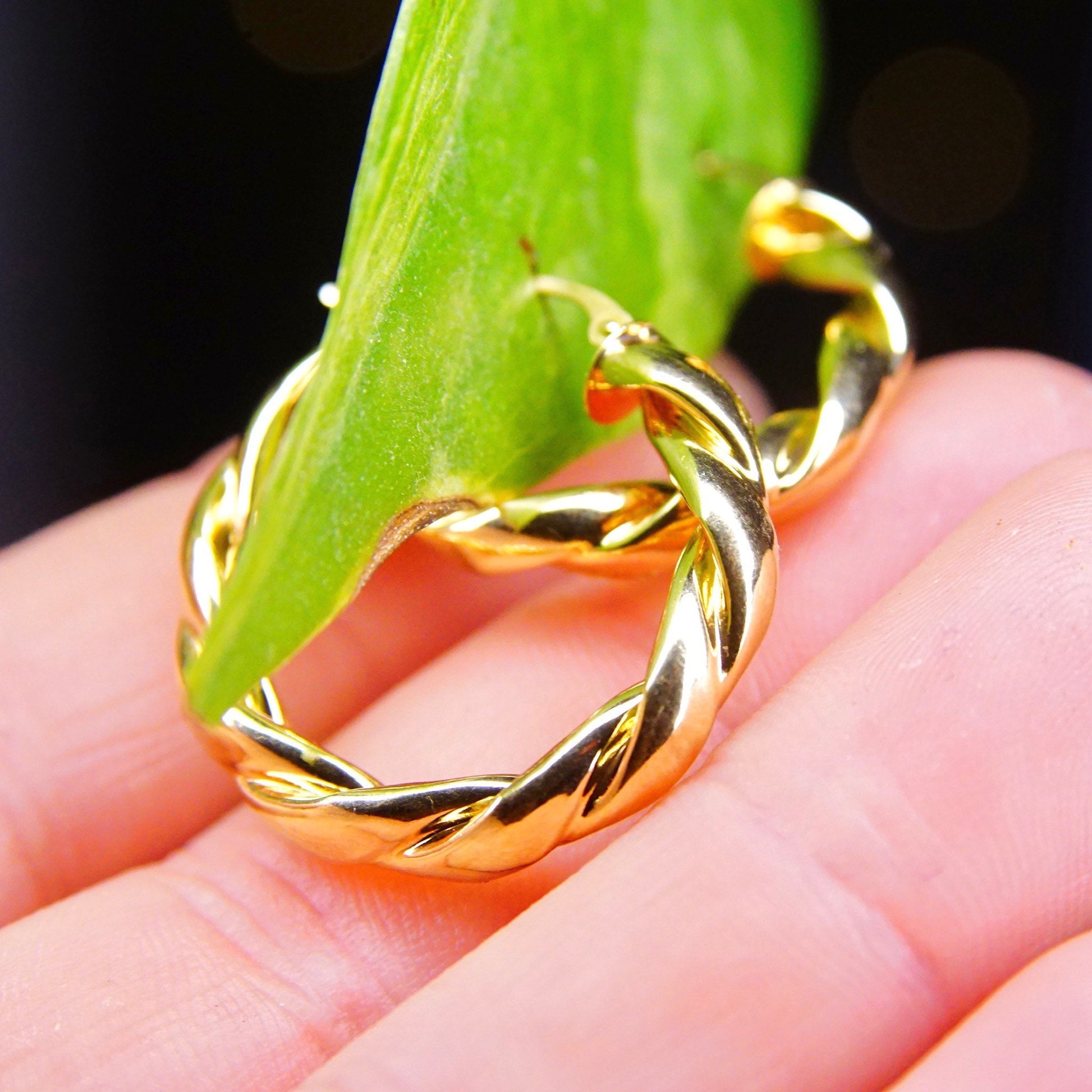 Twisted gold hoop earrings held in hand with green leaf in background