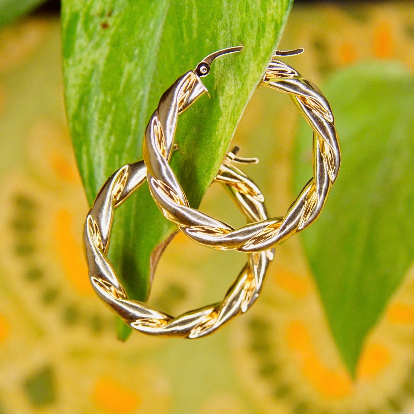 Vintage 14K yellow gold twisted hoop earrings hanging from green leaf, surrounded by orange flowers in soft focus background.