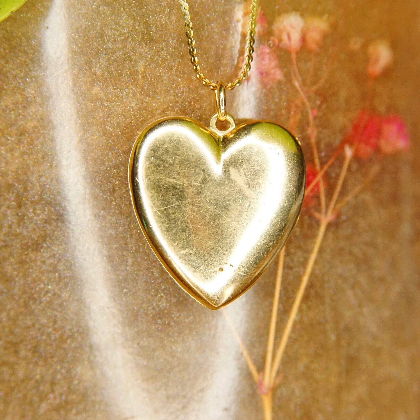 Antique 14K gold heart locket pendant necklace with starburst design and small diamond, hanging in front of shiny gold fabric next to small pink flowers, jewelry photography