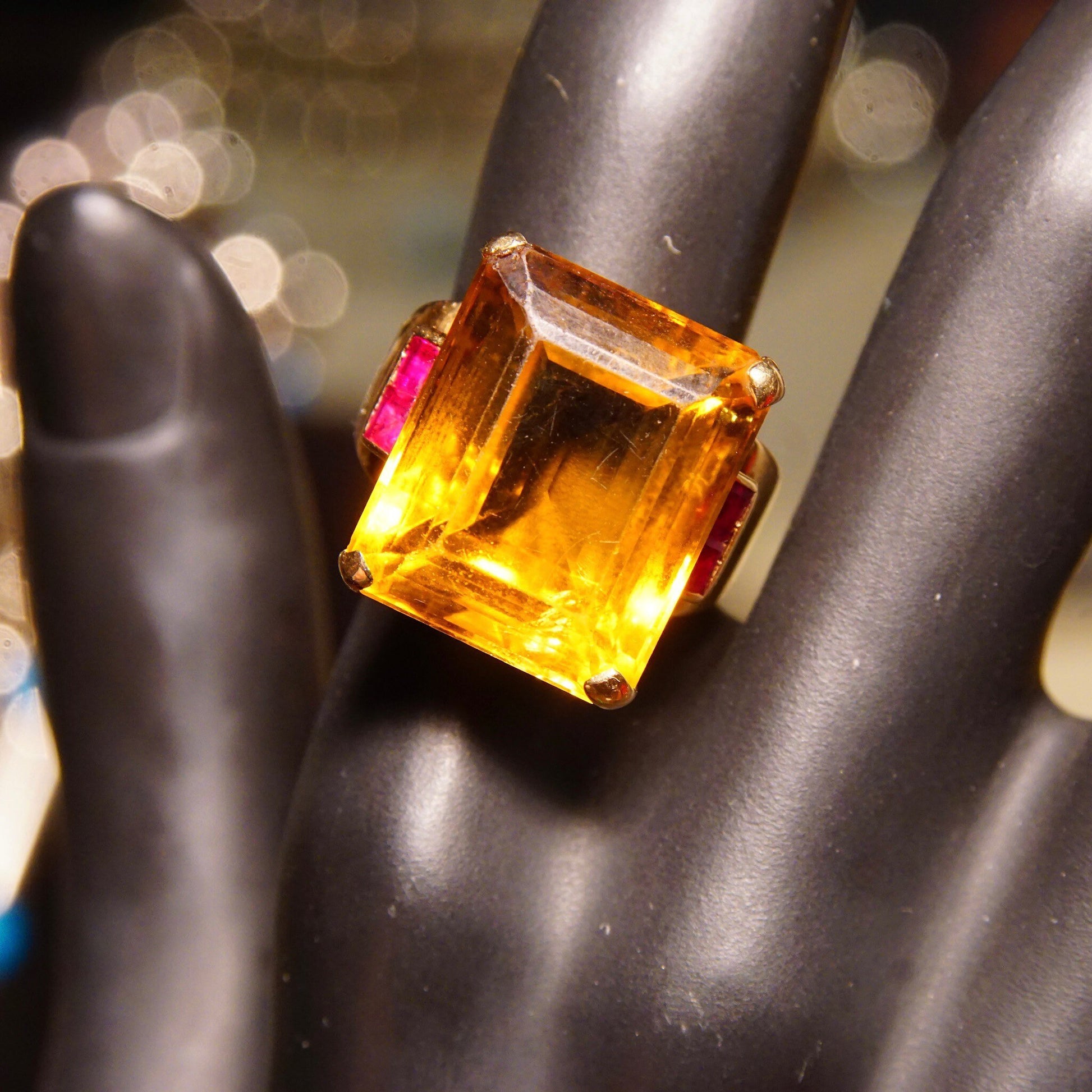 Large emerald-cut citrine gemstone cocktail ring set in 14K yellow gold with ruby accents, estate jewelry, on display held by a mannequin hand.