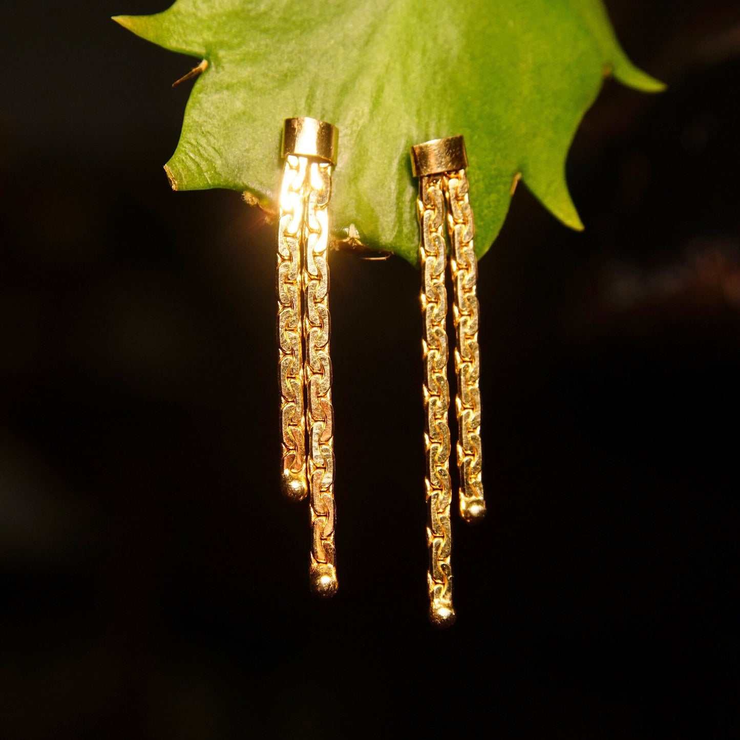 Gold chain tassel earrings with double strands hanging from a leaf, against a black background.