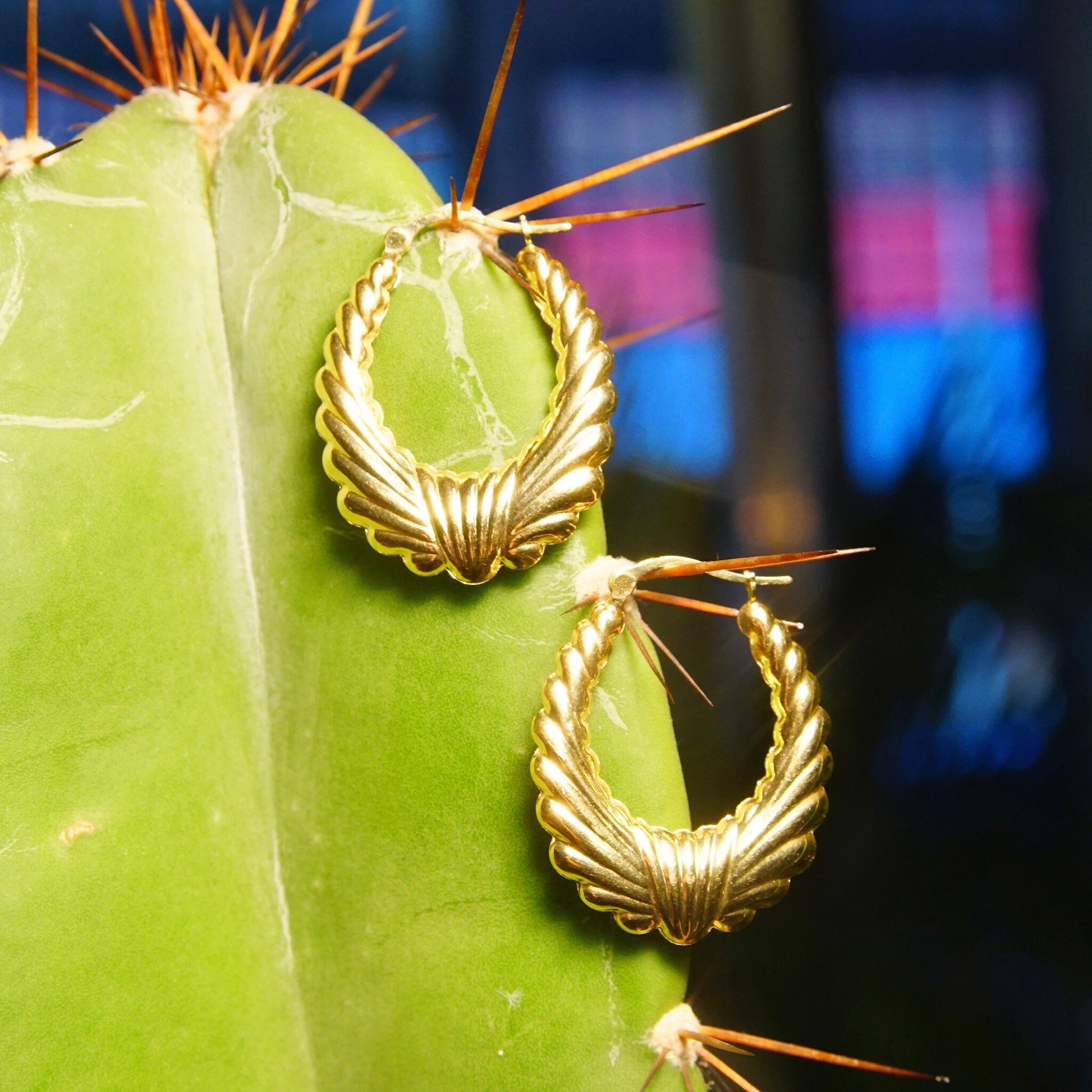 Vintage 14K yellow gold scalloped hoop earrings displayed on a green cactus, with colorful bokeh lights in the background.