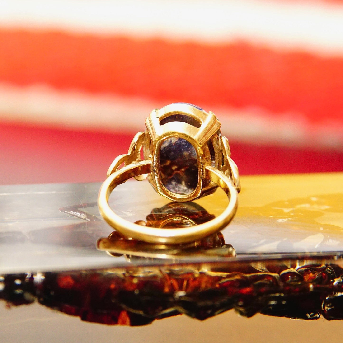 Vintage 10K yellow gold filled ring with dark blue oval lapis lazuli cabochon gemstone featuring golden inclusions, ornate ribbon pattern metal embellishments, size 8 US, against blurred red, white and black background.