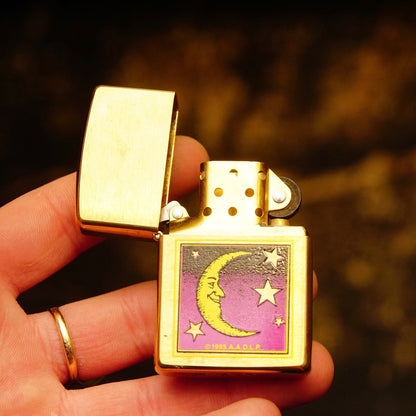 Vintage 1995 AADLP brass Zippo lighter with crescent moon, man in the moon and stars design, made in Bradford, Pennsylvania, USA, measuring 2 1/4 inches, held in hand against blurred background.