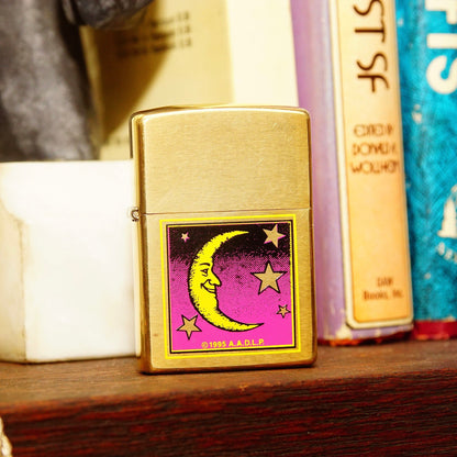 Vintage 1995 AADLP brass Zippo lighter featuring crescent moon, man in the moon, and stars design, made in Bradford, PA, USA, measuring 2 1/4 inches tall, displayed on a wooden shelf with books.