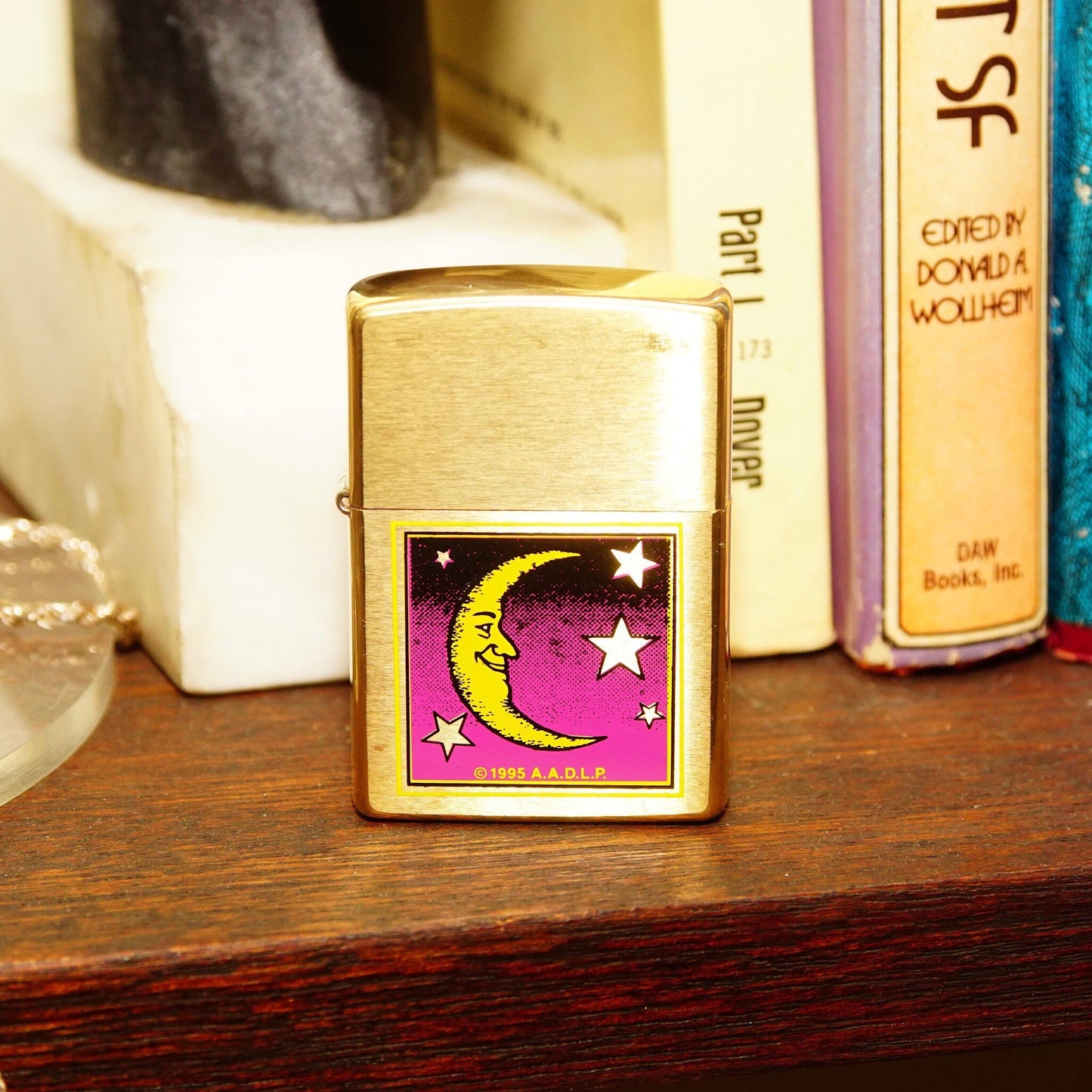 Vintage 1995 AADLP brass Zippo lighter featuring colorful moon and stars design, made in Bradford, Pennsylvania, USA, measuring 2 1/4 inches tall, displayed next to books on a wooden surface.