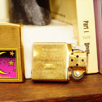 Vintage brass Zippo lighter from 1995 featuring a crescent moon design with a man's face and stars, made in Bradford, Pennsylvania, USA, measuring 2 1/4 inches tall, displayed next to matchbooks and books on a wooden surface.