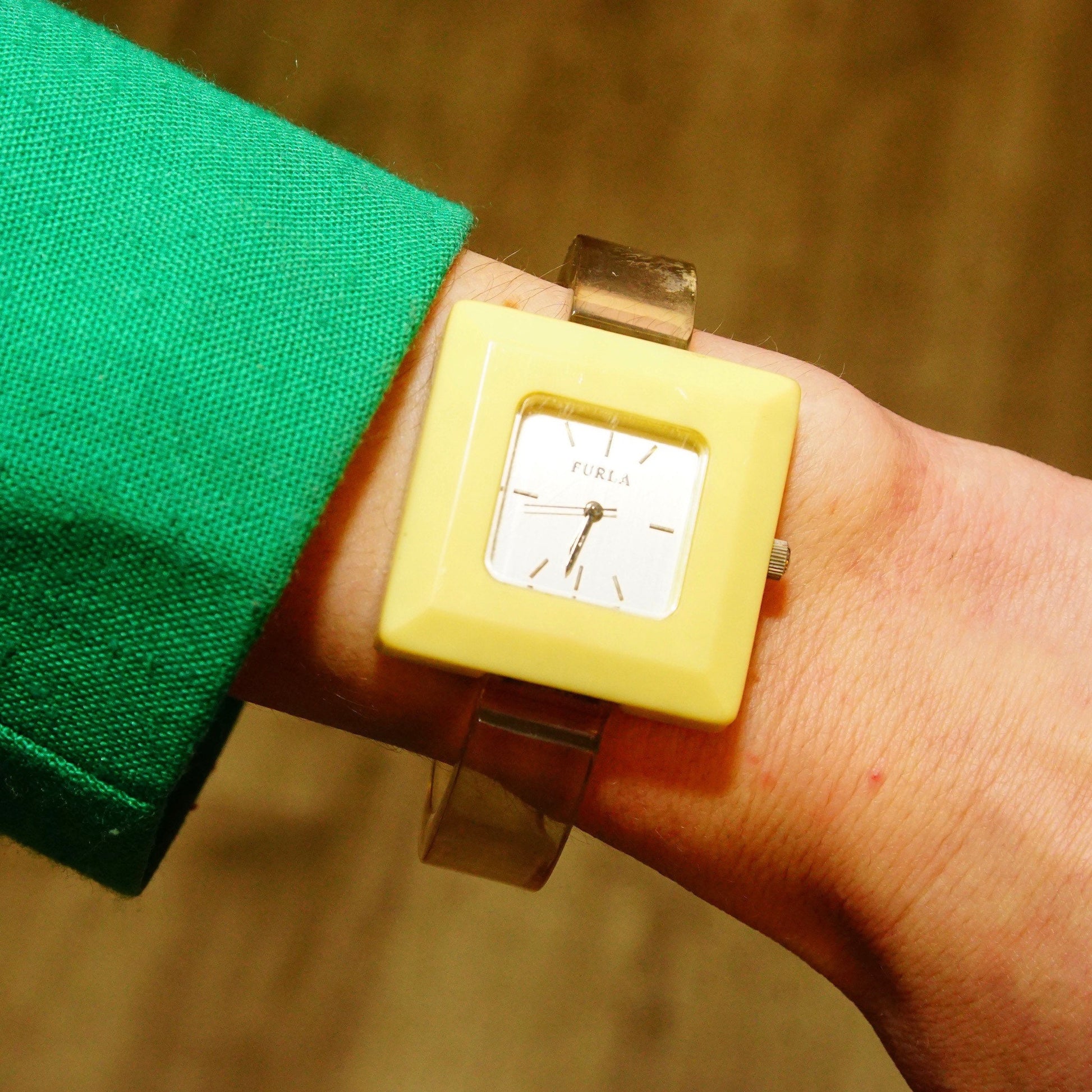 Vintage Furla cuff wristwatch with square beige plastic case and reflective dial on a person's wrist wearing a green sleeve, crafting a chunky, retro accessory look.