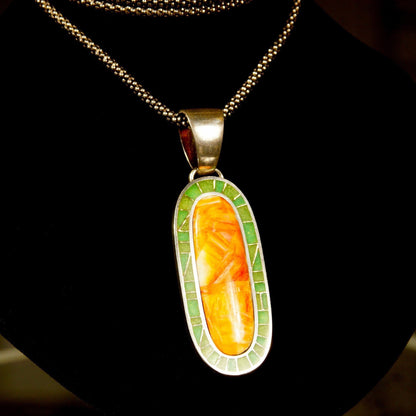 Vintage 925 sterling silver pendant necklace featuring a long oval design with orange carnelian agate and green aventurine inlay in a mosaic pattern, suspended from a large silver bail on a chain against a black background.