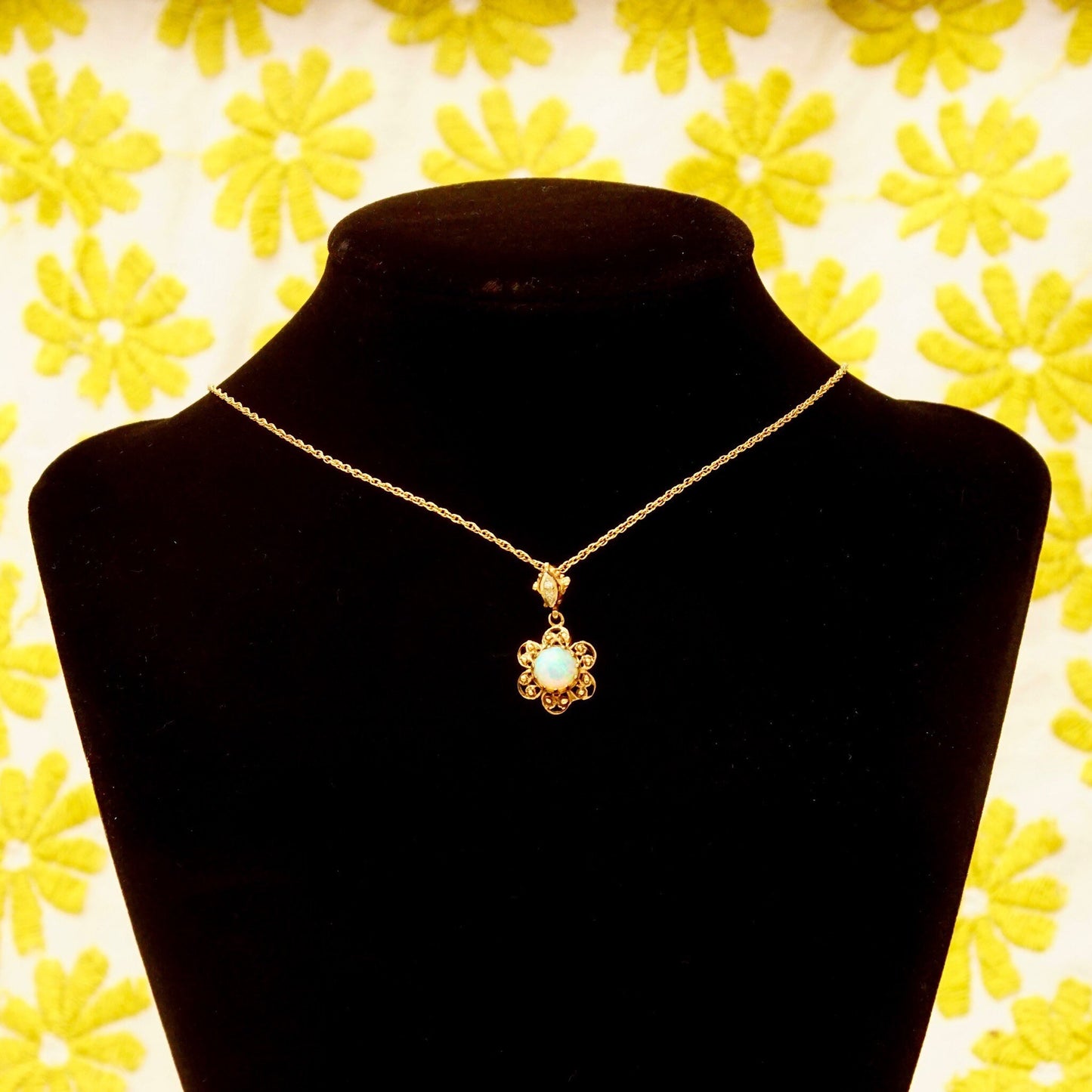 14K gold filigree opal flower pendant necklace with diamond accent bail on black velvet jewelry display stand against yellow floral wallpaper background.