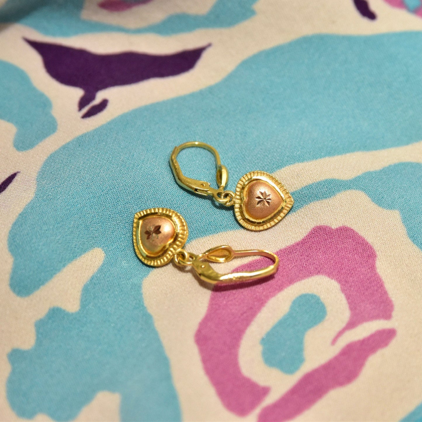 Vintage 14K yellow and rose gold heart dangle earrings with diamond-cut accents on teal and purple abstract patterned background