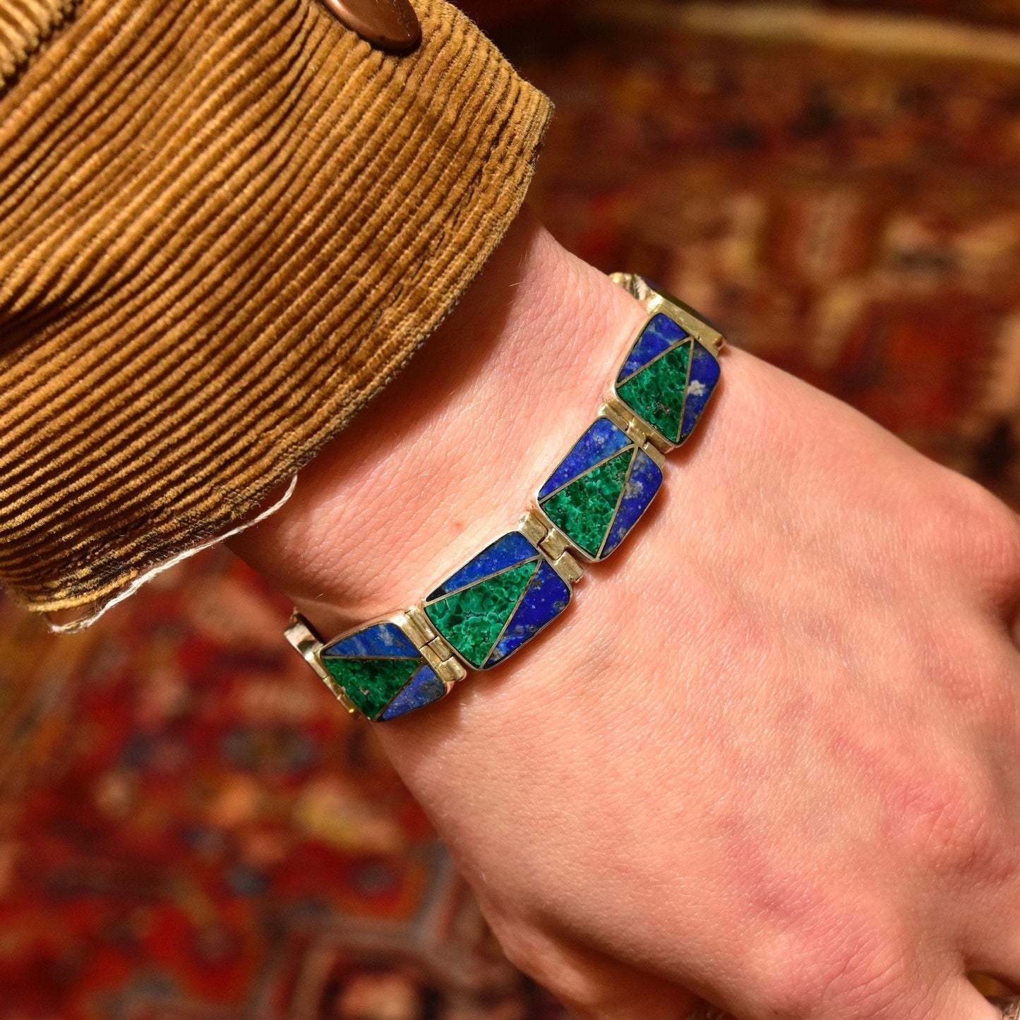 Vintage sterling silver link bracelet with geometric malachite and lapis lazuli inlay on a wrist, set against a colorful patterned background.