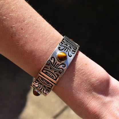 Vintage TAXCO sterling silver link bracelet with tiger's eye stone and tribal motifs, worn on a person's wrist. The heavy silver cuff-style bangle has a security chain clasp and measures approximately 7 1/2 inches in length.