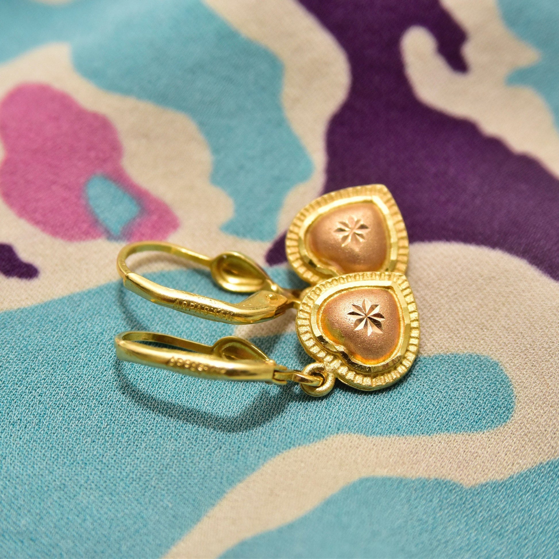 Pair of 14K yellow gold dangle earrings with diamond-cut rose gold heart charms on a colorful fabric background