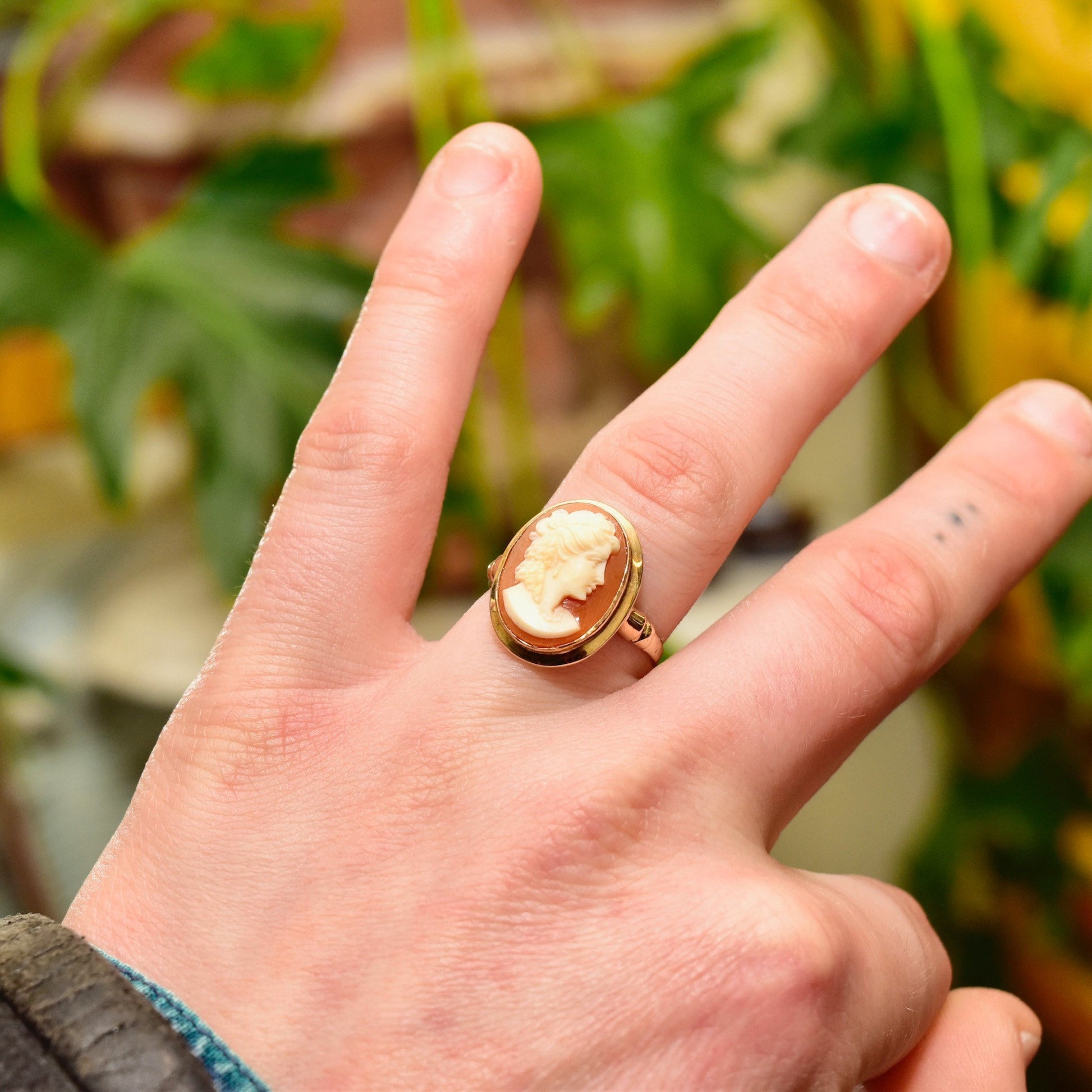 Alt text: Hand wearing vintage 14K gold cameo ring with shell carving, on blurred background of green leaves