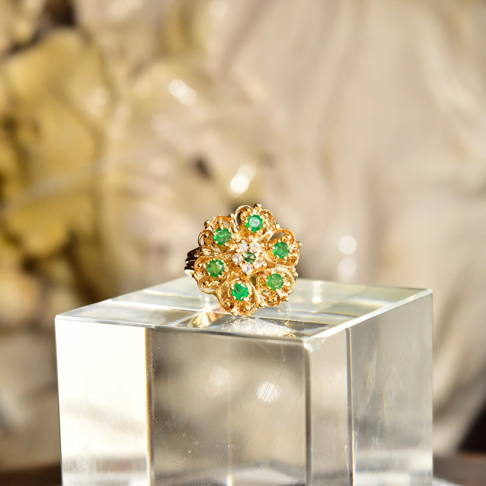 14K yellow gold emerald and diamond cluster cocktail ring in a floral dome design, displayed on a clear glass pedestal against a textured beige background.