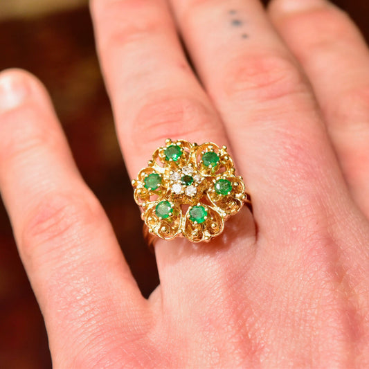 14K yellow gold emerald and diamond cluster dome cocktail ring on a hand, featuring a floral design set with round green emeralds surrounded by sparkling diamonds, ring size 7 1/2 US.