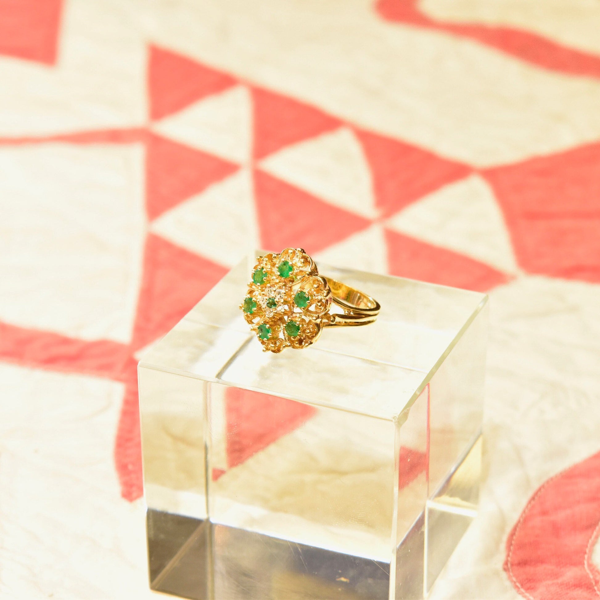 14K yellow gold emerald and diamond cluster dome cocktail ring, flower design, size 7.5 US, displayed on geometric marble pedestal against red and cream geometric background.