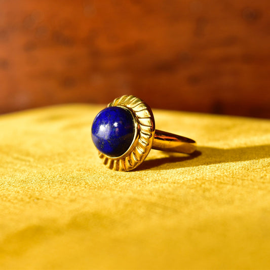 14K yellow gold cocktail ring featuring a round cobalt blue lapis lazuli cabochon gemstone set in a fluted bezel setting, displayed on a textured golden fabric background.