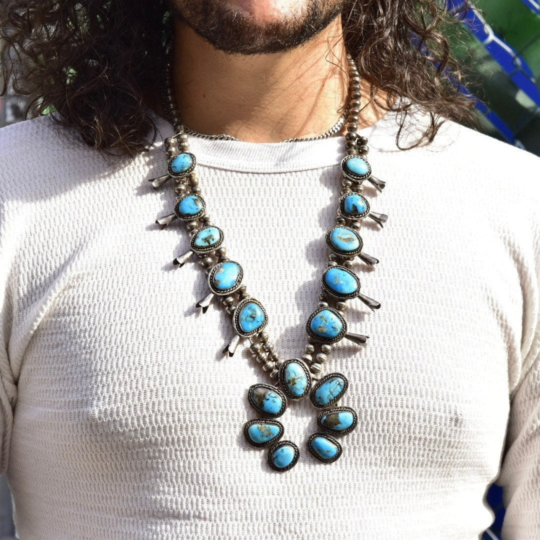 Vintage Navajo squash blossom necklace with natural blue turquoise stones set in coin silver, worn by a person with long curly hair and a white sweater, showing the intricate design and length of this traditional Native American old pawn jewelry piece.