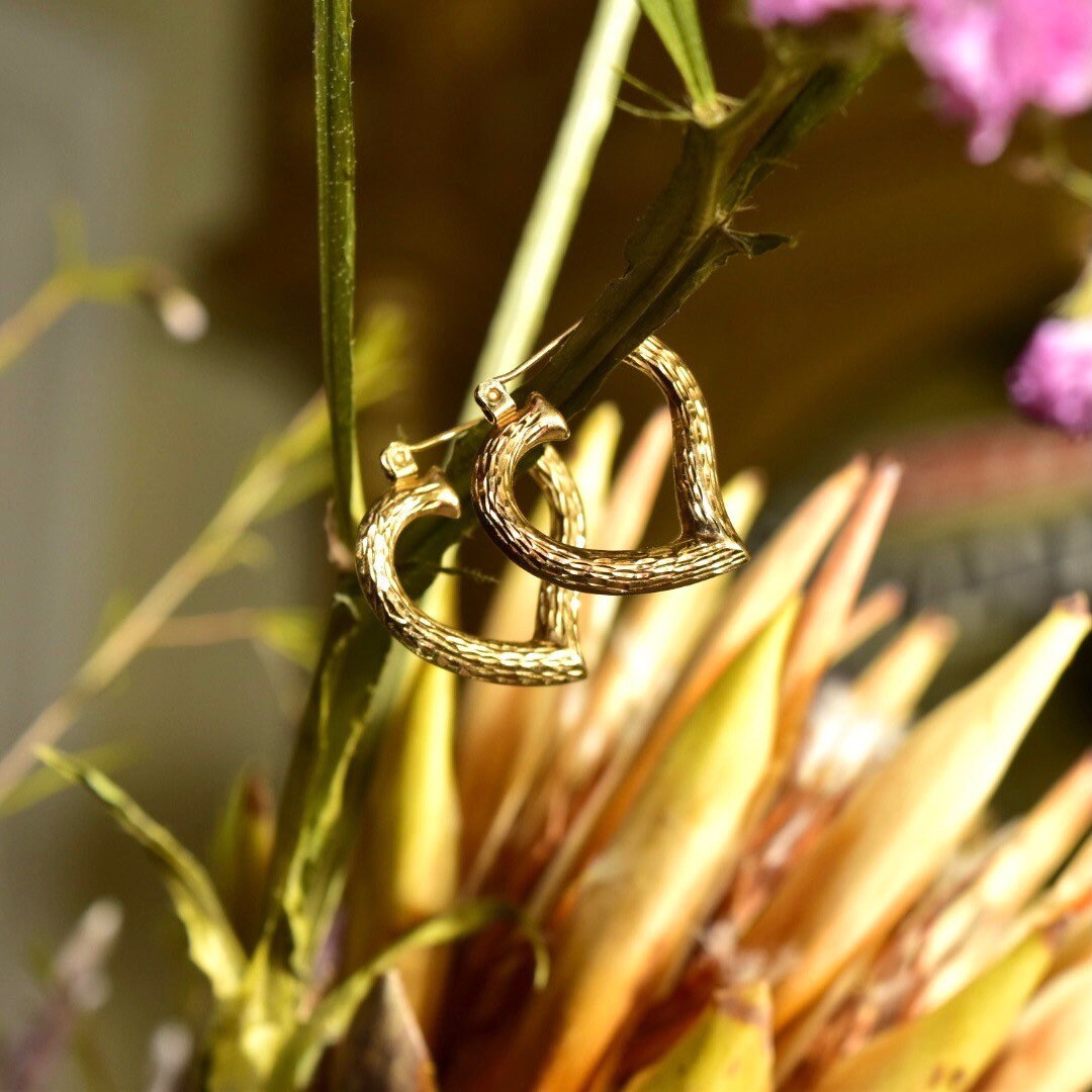 Vintage 14K yellow gold hammered heart hoop earrings hanging from plant stem, showing intricate textured surface and open heart design against blurred floral background.