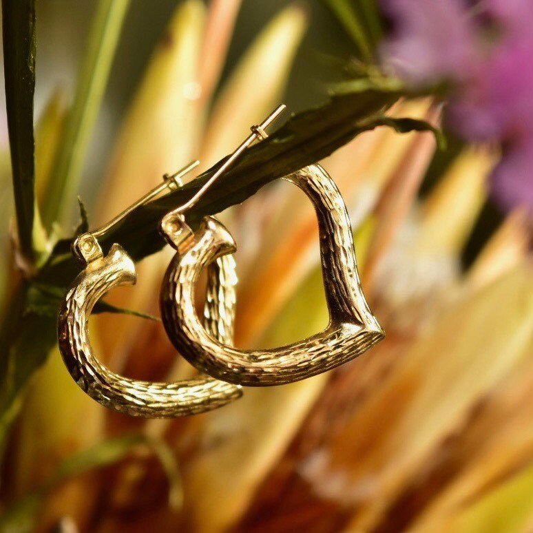 Vintage 14K yellow gold hammered heart hoop earrings hanging from plant stem, showing textured surface and heart-shaped design, 22mm in diameter.