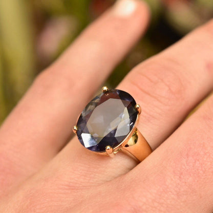 Vintage gold plated sterling silver ring with a large oval-cut blue simulated gemstone on a hand, showing the ring from the side against a blurred natural green background.