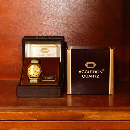 Vintage 1988 Bulova Accutron gold tone Swiss quartz day/date wristwatch with original box and papers displayed on wooden surface.