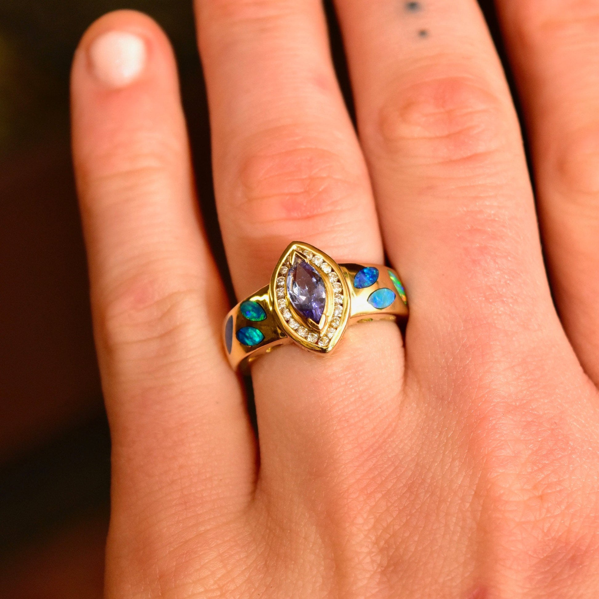 14K gold marquise tanzanite ring with diamond halo and blue opal inlay accents, worn on a finger against a plain background.