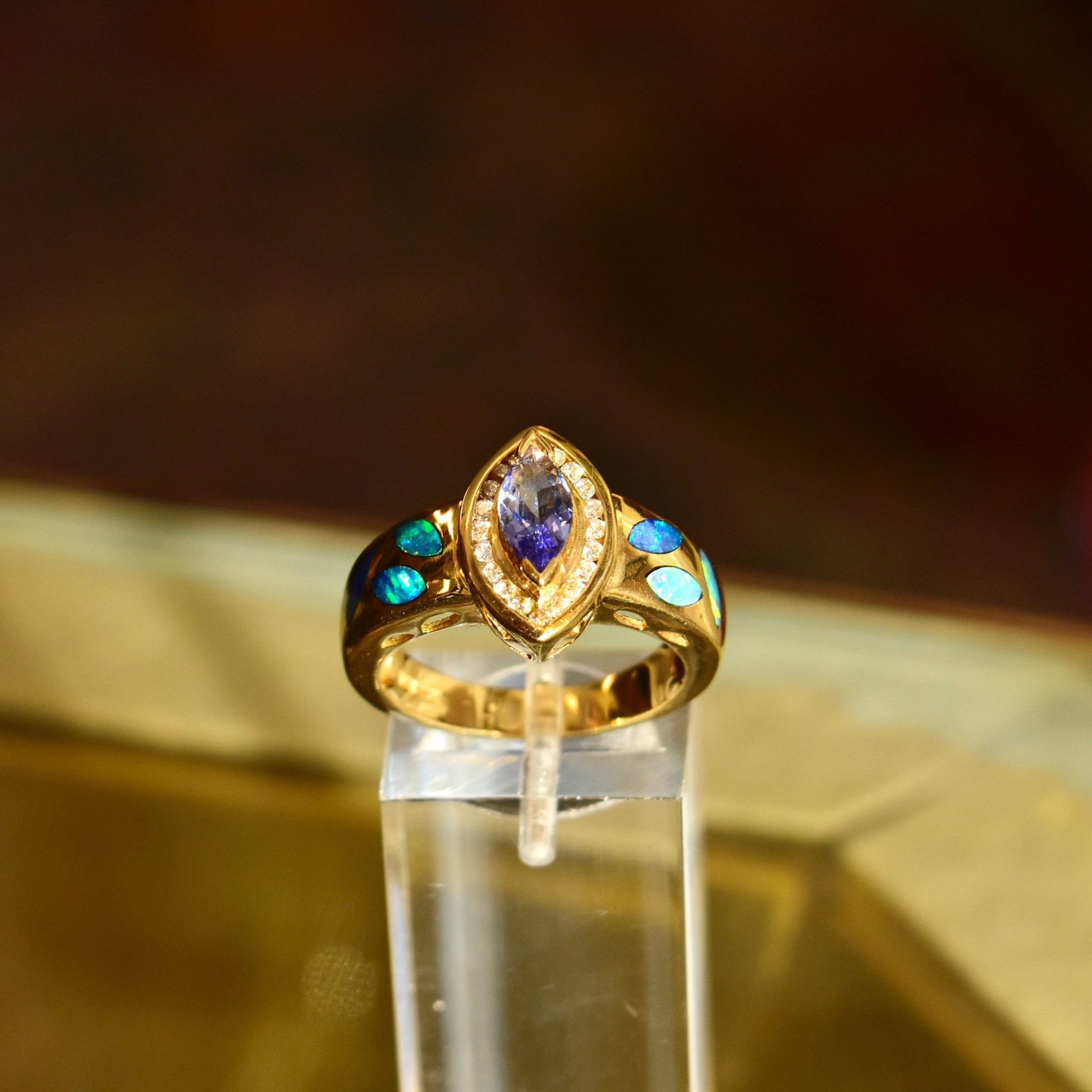 14K gold cocktail ring featuring a marquise-cut tanzanite center stone accented by a diamond halo and vibrant blue opal inlays, shown on a neutral fabric background.