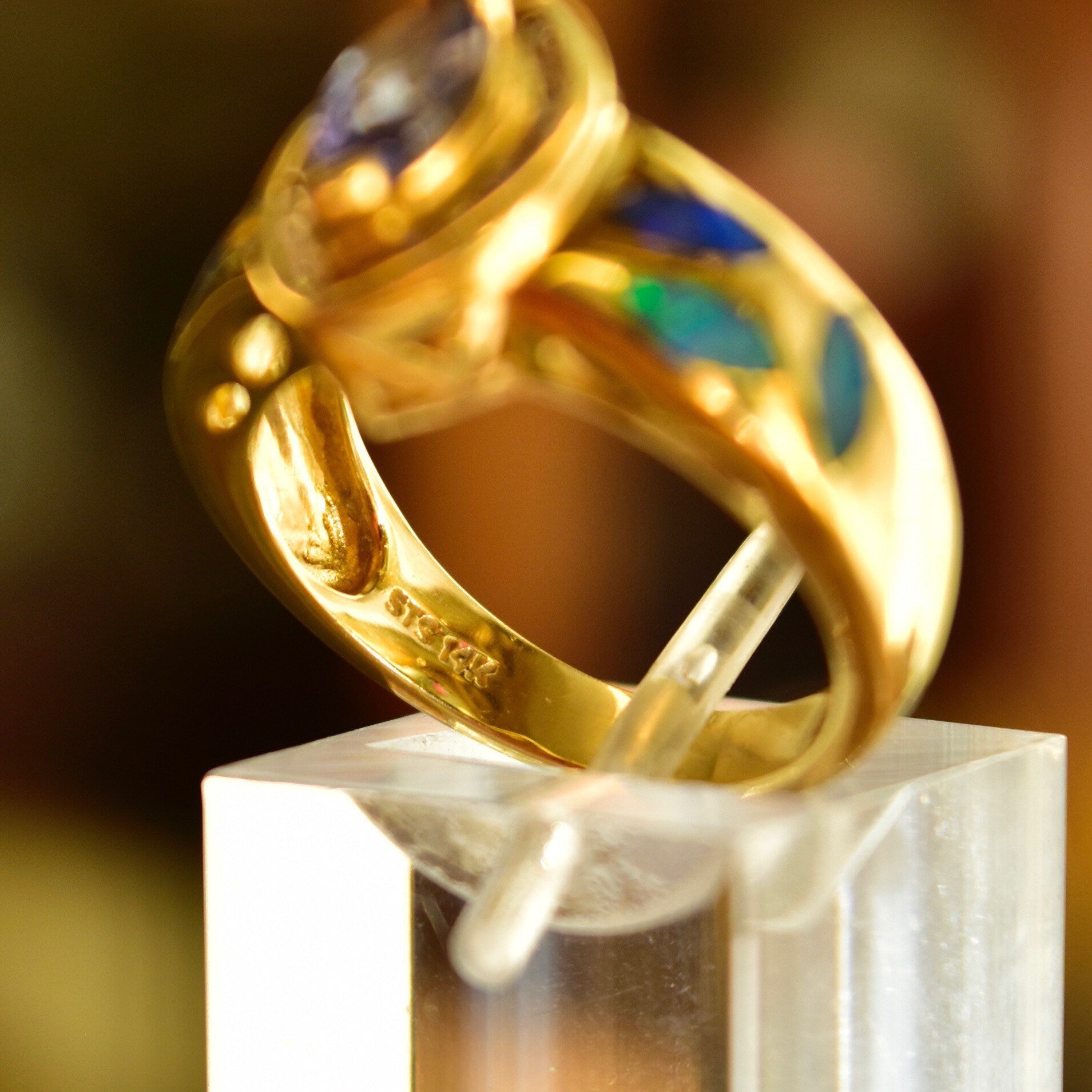 Golden ring with marquise-cut tanzanite center stone surrounded by diamond halo and blue opal inlay, displayed on white jewelry stand against blurred brown background