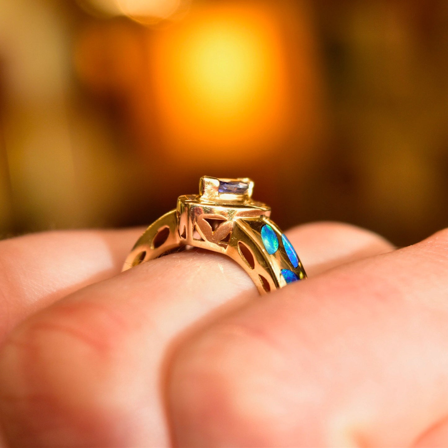 14K gold ring with marquise tanzanite center stone surrounded by diamond halo, featuring blue opal inlay on the band, resting on a person's hand, illuminated by warm lighting in a close-up view