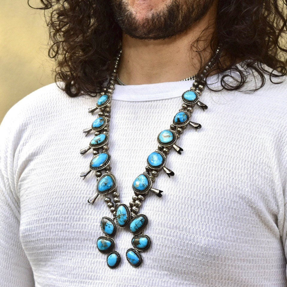 Vintage Navajo squash blossom necklace featuring natural blue turquoise stones set in coin silver, worn by a person with curly hair and a white top. The Native American old pawn jewelry piece measures 29 inches long.