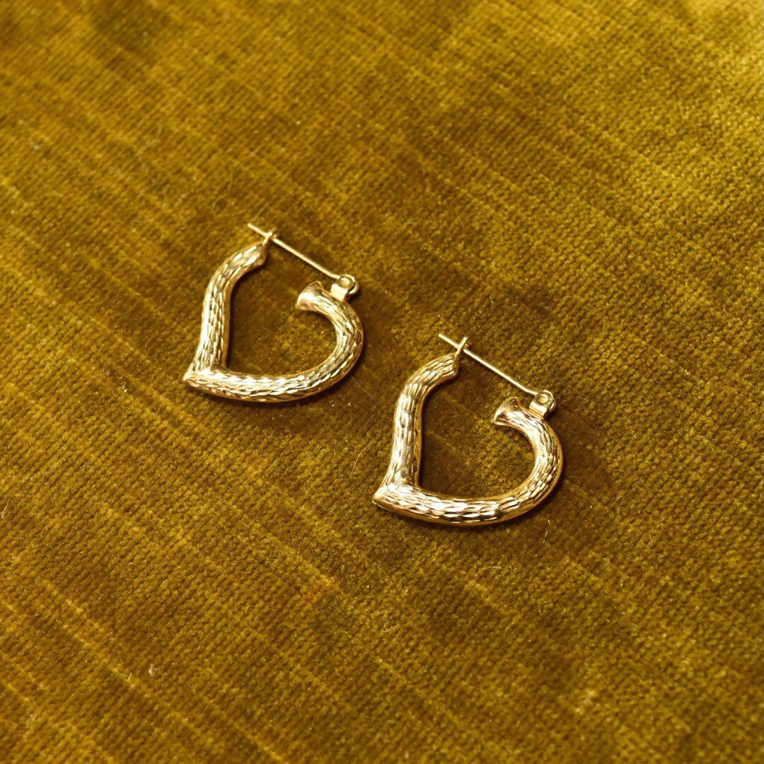 Vintage 14K yellow gold hammered textured heart hoop earrings, 22mm in size, laying on golden fabric background.