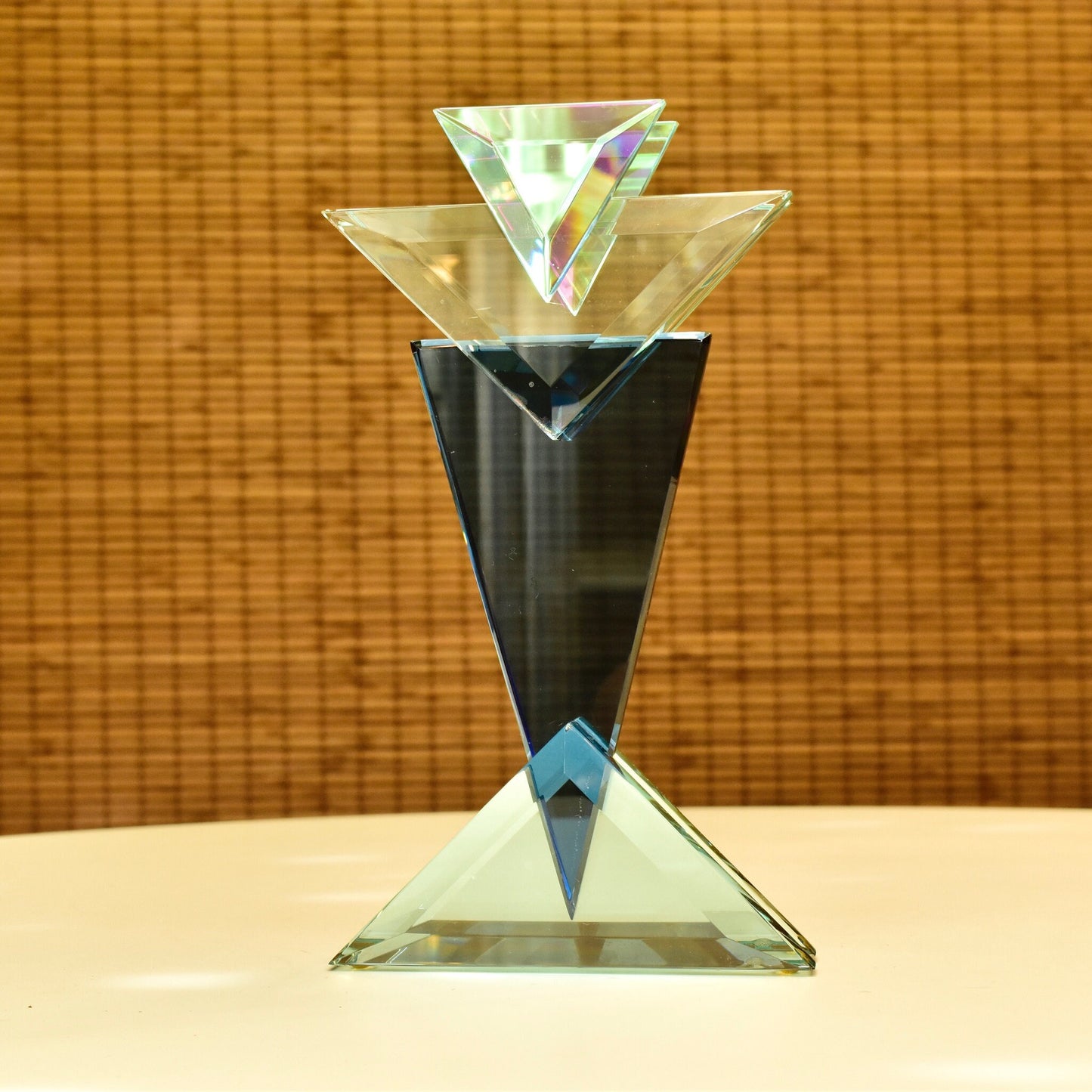 Vintage 1989 signed American studio glass geometric sculpture by Stephen Jon Clements featuring stacked triangular prisms in iridescent blue, green and clear glass on a clear triangular base, measuring 10 1/8 inches tall, displayed on a woven rattan surface.