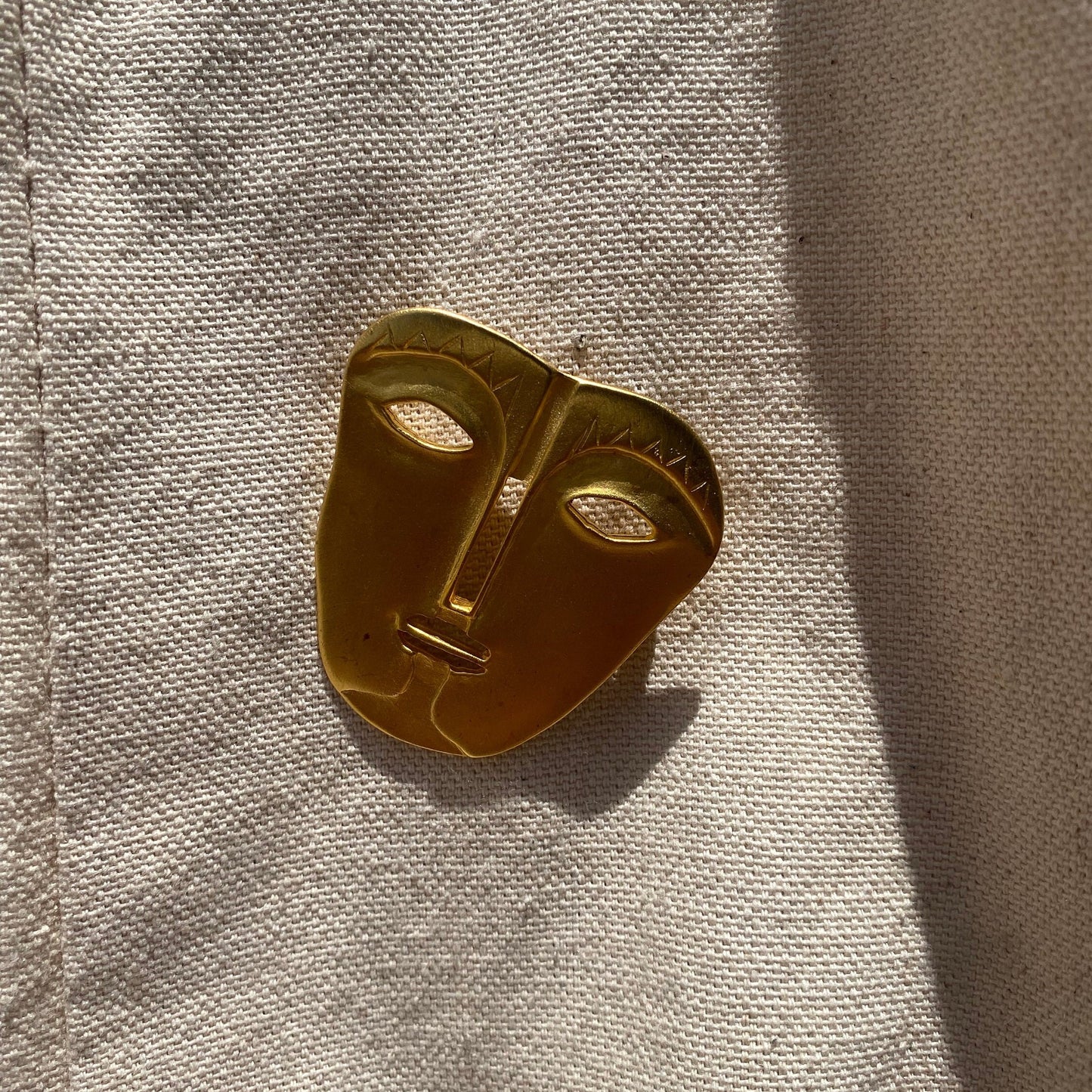 Vintage gold-toned abstract tribal face mask brooch pin with engraved details on textured fabric background, 45 mm unique modernist jacket pin accessory