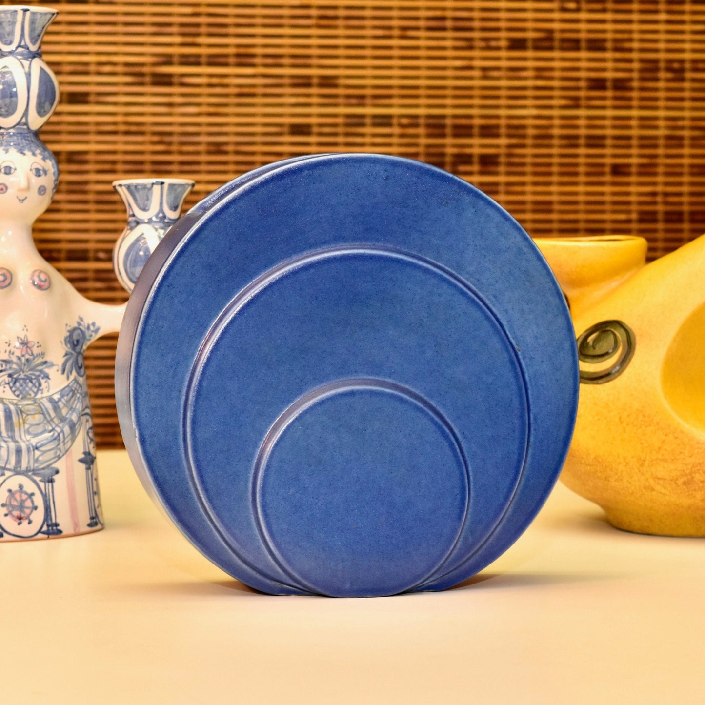 Vintage blue Art Deco ceramic vase with circular disk design by Trenton Potteries, designed by G. McStay Jackson. The modernist sculptural vase features concentric rings in a deep blue glaze and measures 8 1/2 inches tall. Displayed with other decorative ceramic objects against a textured background.