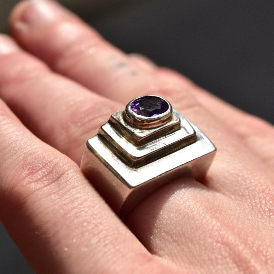 Vintage modernist chunky sterling silver ring with an oval cut amethyst gemstone set in a textured, stepped dome design, shown on a hand. The ring is size 6 1/2 US.