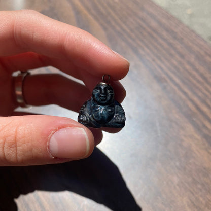 Vintage carved black stone Buddha charm with silver loop held between fingers, showing the small, cute Buddha figure in detail against a wooden surface background.