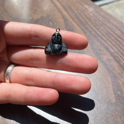 Vintage carved black stone Buddha charm pendant held in palm, showing small size and intricate details of the seated Buddha figure. The charm has a silver loop at the top for attaching to jewelry.