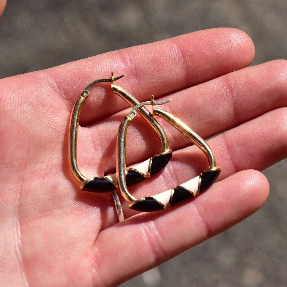 14K gold triangle hoop earrings with black and beige enamel designs held in palm of hand
