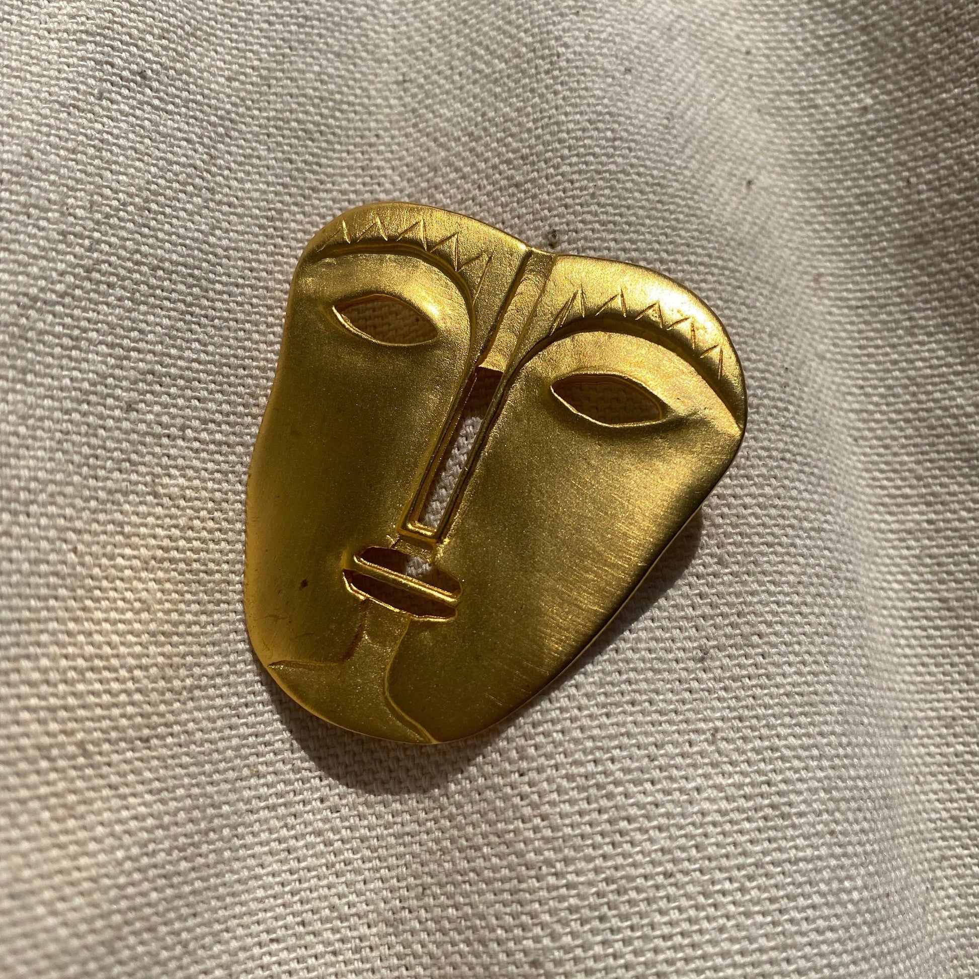 Gold abstract tribal face mask brooch pin with engraved details on textured fabric background