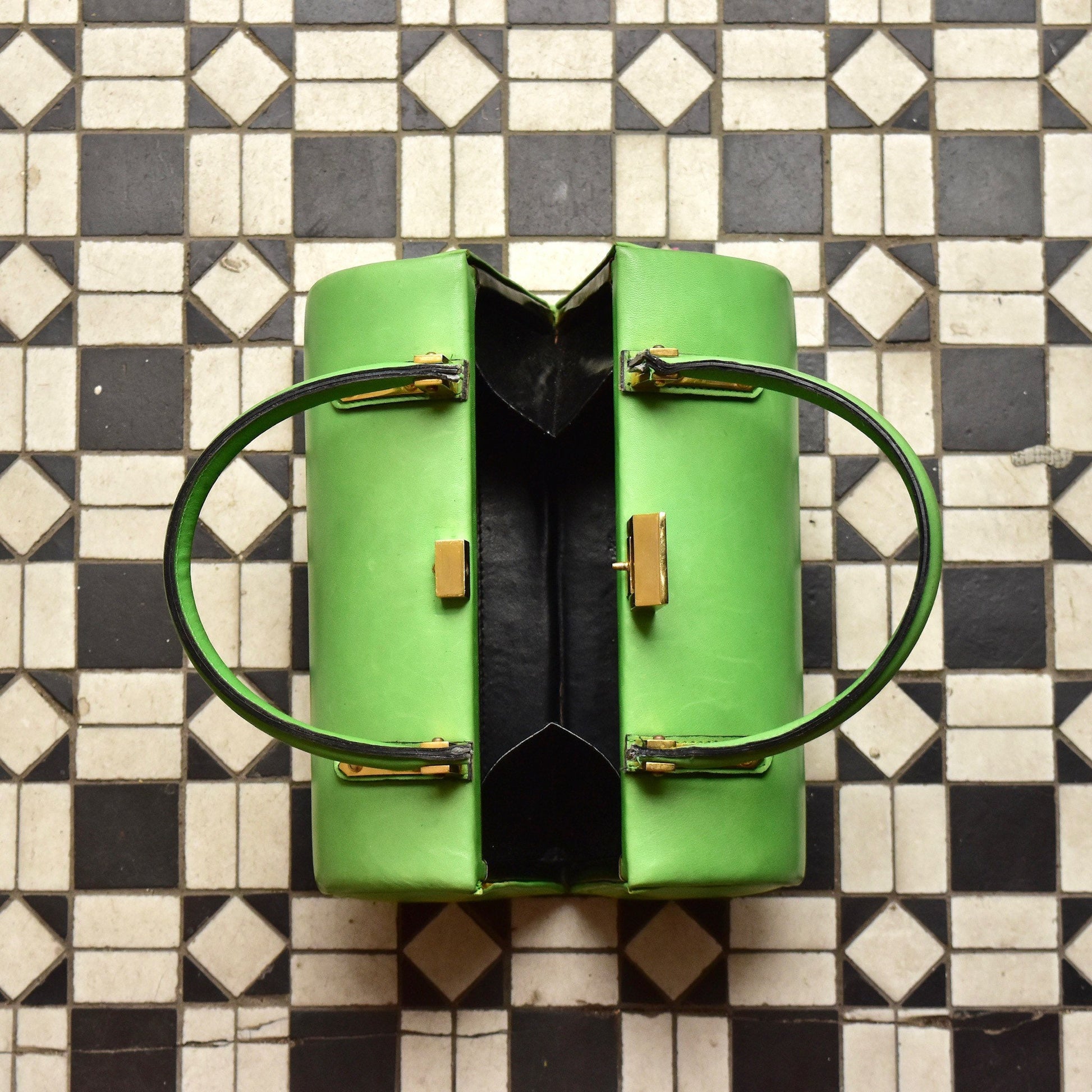 Mod TANO of Madrid lime green leather barrel box purse with black trim, brass hardware, and top handles, placed on a black and white tile floor.