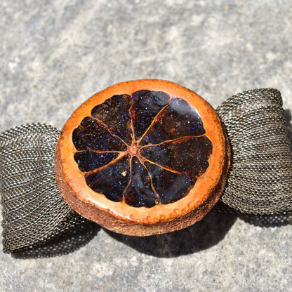 Preserved orange slice snap bracelet with sparkly iridescent purple glaze on a silver mesh adjustable band, shown on gray stone background.