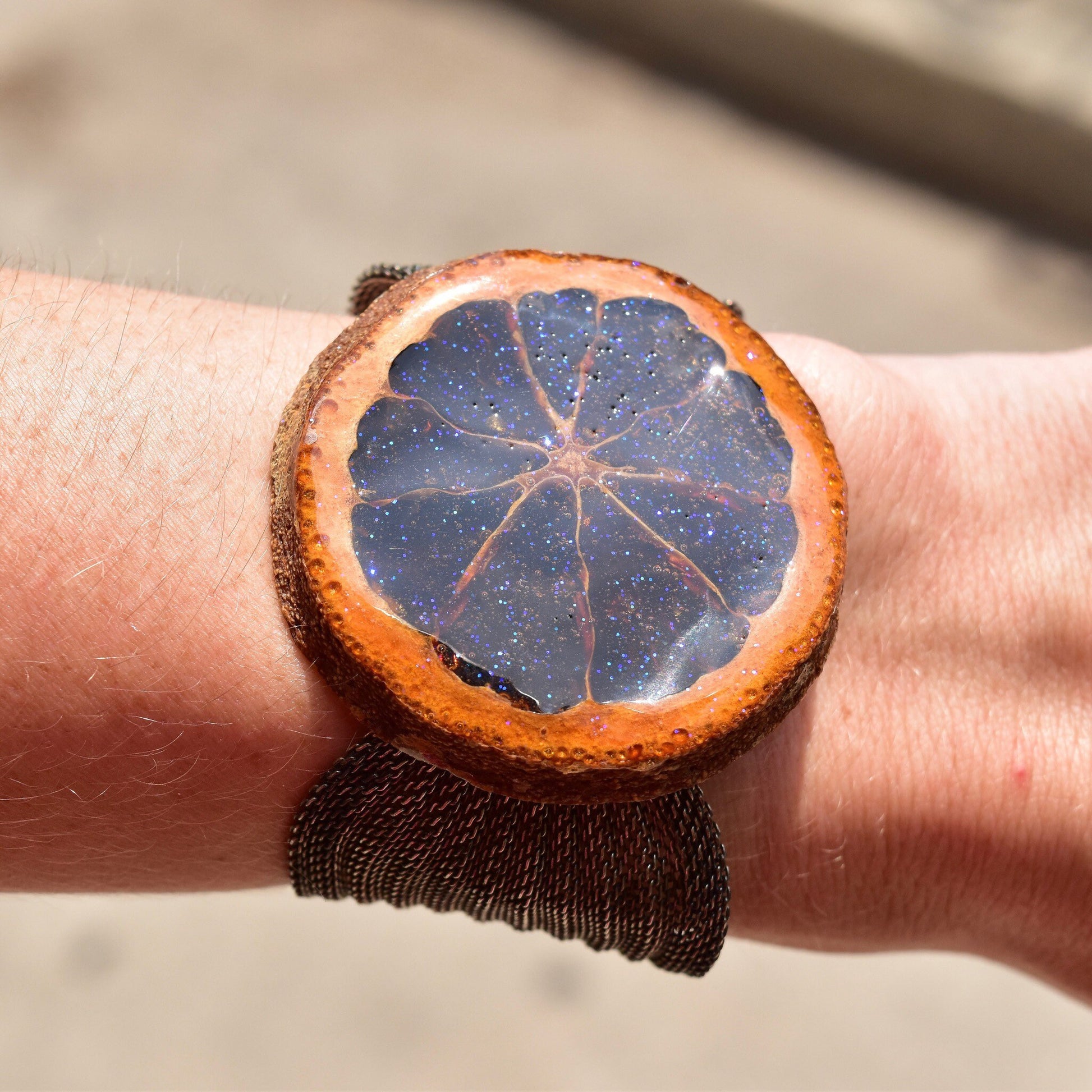 Preserved orange slice bracelet with iridescent purple glaze on silver mesh band, showing cross-section of citrus against wrist