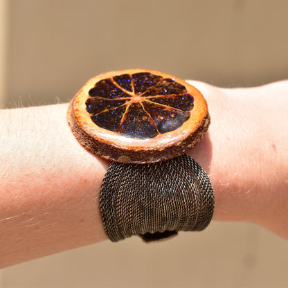 Unique sparkly preserved orange slice snap cuff bracelet with silver mesh band and iridescent purple glaze, adjustable length from 6 1/2 to 7 1/4 inches, shown on a wrist.