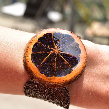 Unique sparkly preserved orange slice bracelet with iridescent purple glaze on an adjustable silver mesh band, shown worn on a wrist.