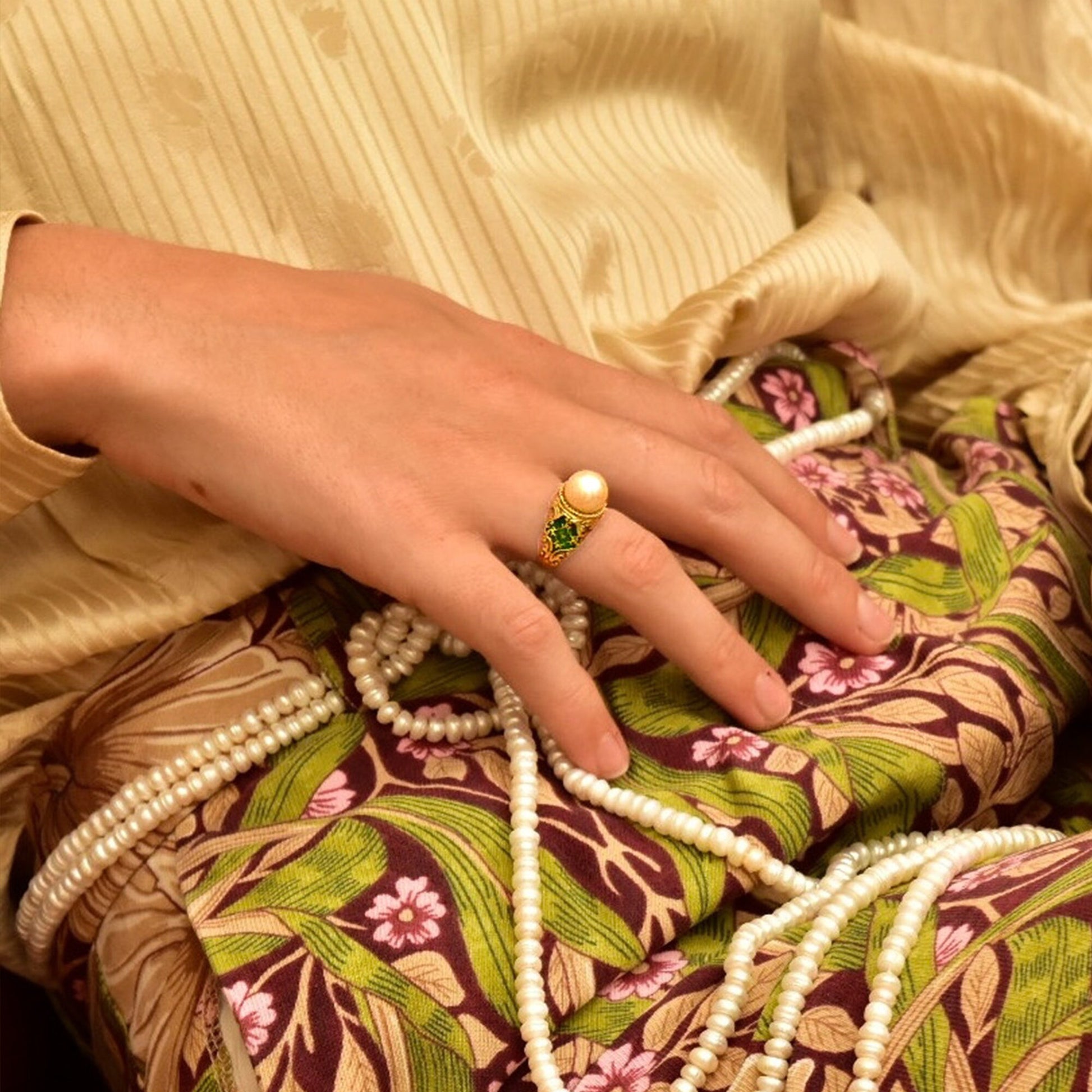 A woman's hand wearing an ornate vintage pearl solitaire ring with emerald green guilloche enamel designs resting on a floral patterned fabric.