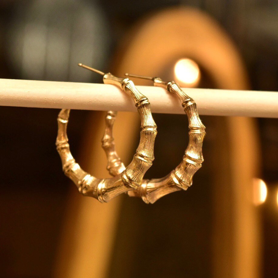14K yellow gold bamboo hoop earrings with stud post closures, hanging from a wooden dowel, slightly out of focus background.